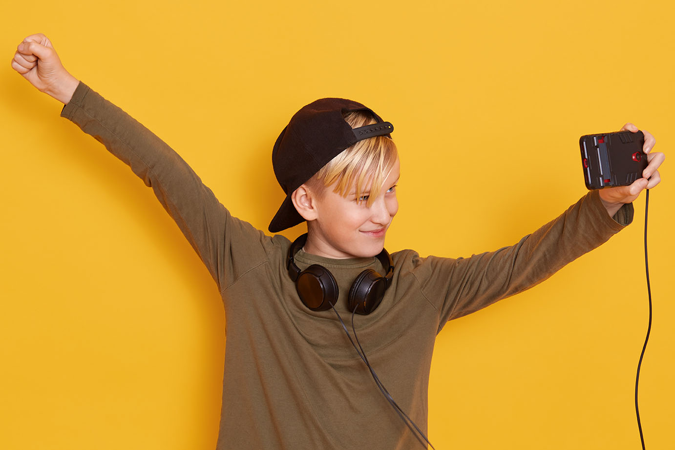 Close up portrait of little boy holding his modern cellphone and watching video,using wireless Internet and headphones, child dresses green hoody and blackcap, posing isoalted over yellow background.